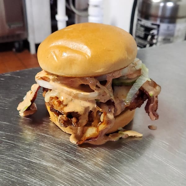 A Brake Burger Garage burger with sauce, onions, and pickles overflowing onto a metal surface.